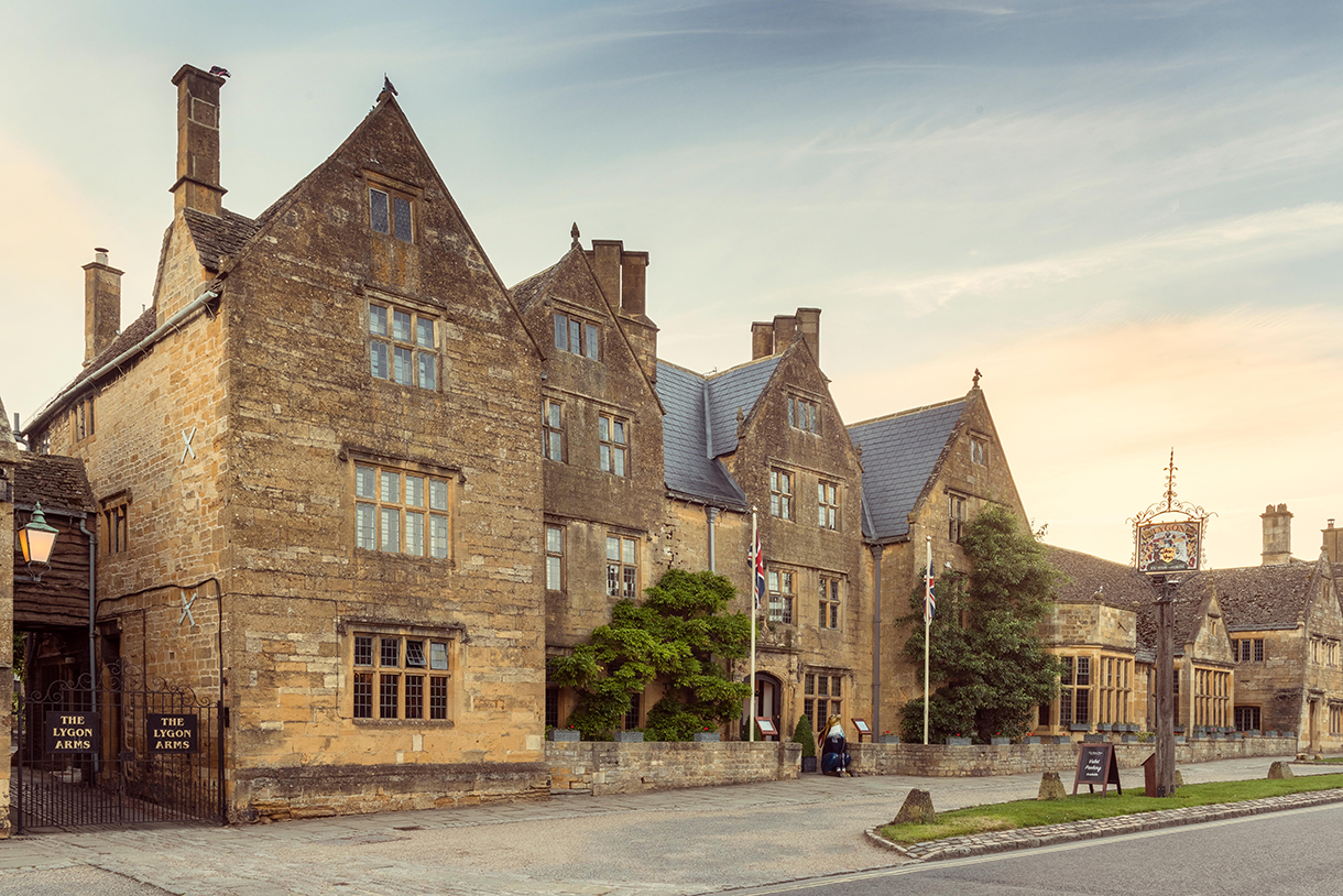 Exterior of Lygon Arms Hotel, a large historic building with a brick facade and black roof, surrounded by green trees and bushes.